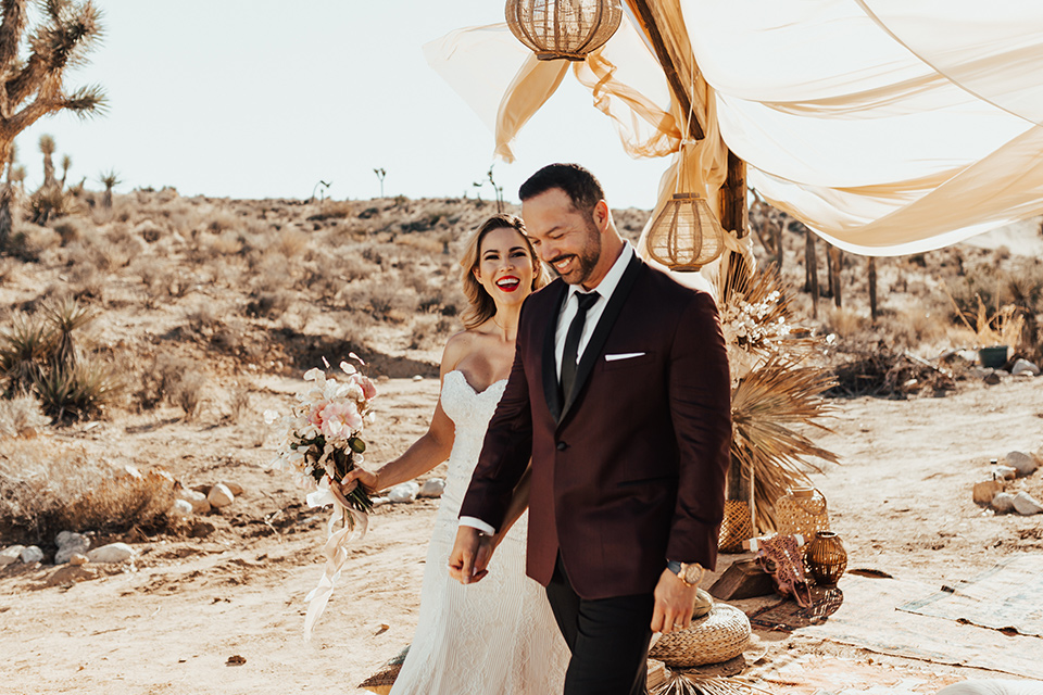 Bohemian-Desert-Shoot-bride-and-groom-walking-towards-camera-bride-in-a-formfitting-gown-with-lace-detailing-and-a-sweetheart-neckline-groom-in-a-burgundy-shawl-lapel-tuxedo-with-a-black-long-tie