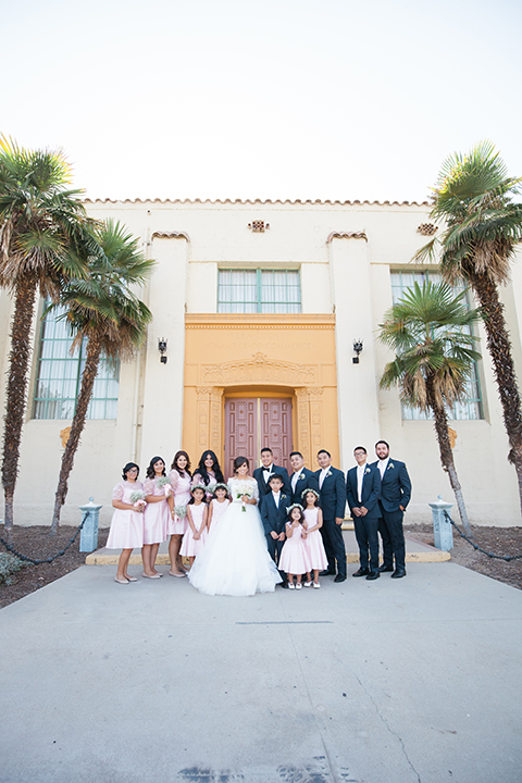 Elements-Venue-presents-wedding-bridal-party-outside-bridesmaids-in-pink-gowns-groomsmen-in-navy-suits-with-pink-long-ties-bride-is-in-a-layered-tulle-ball-gown-with-a-high-neckline-and-quarter-sleeves-groom-in-a-navy-blue-shawl-lapel-tuxedo-with-a-black-bow-tie