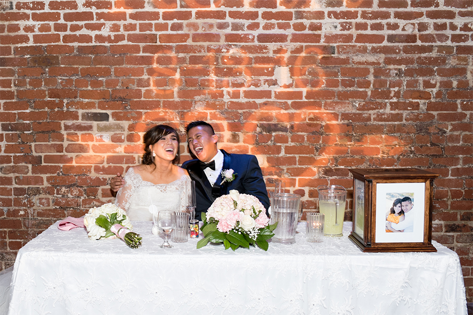 Elements-Venue-presents-wedding-bride-and-groom-sitting-at-sweetheart-table-bride-is-in-a-layered-tulle-ball-gown-with-a-high-neckline-and-quarter-sleeves-groom-in-a-navy-blue-shawl-lapel-tuxedo-with-a-black-bow-tie