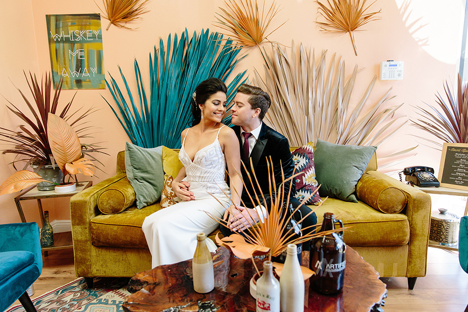 bride and groom sitting on yellow couch bride wore a form fitting white gown with lace detailing and a deep v neckline the groom is in a black velvet tuxedo with a peak lapel and burgundy long tie