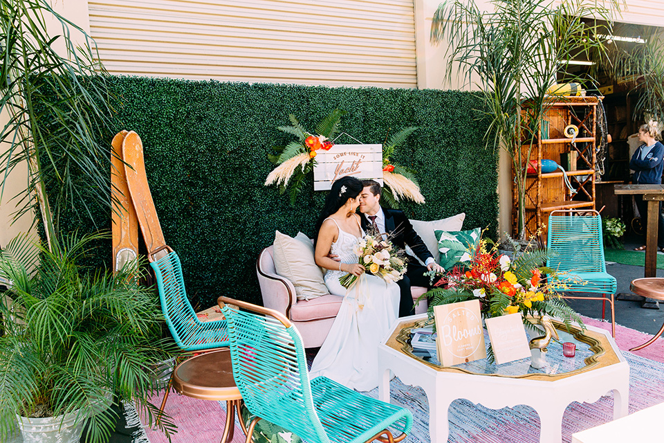 bride and groom sitting on outside furniture long shot the bride wore a from fitting white gown with lace detailing and a deep v neckline the groom is in a black velvet tuxedo with a peak lapel and burgundy long tie