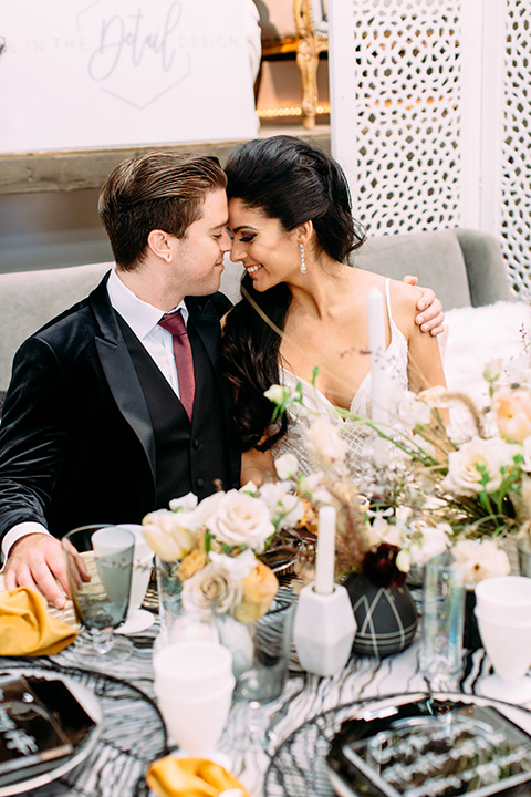 bride and groom touching heads at sweetheart table the bride is wearing a white lace form fitting gown with a deep v neckline and the groom wore a black velvet tuxedo with a burgundy long tie