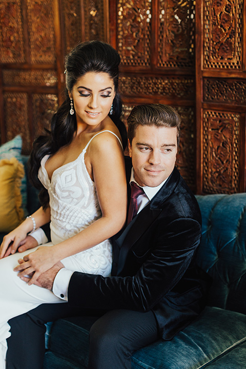 bride sitting on grooms lap with the bride wearing a form fitting white gown with lace detailing and a deep v neckline the groom wore a black velvet tuxedo with a peak lapel and burgundy long tie