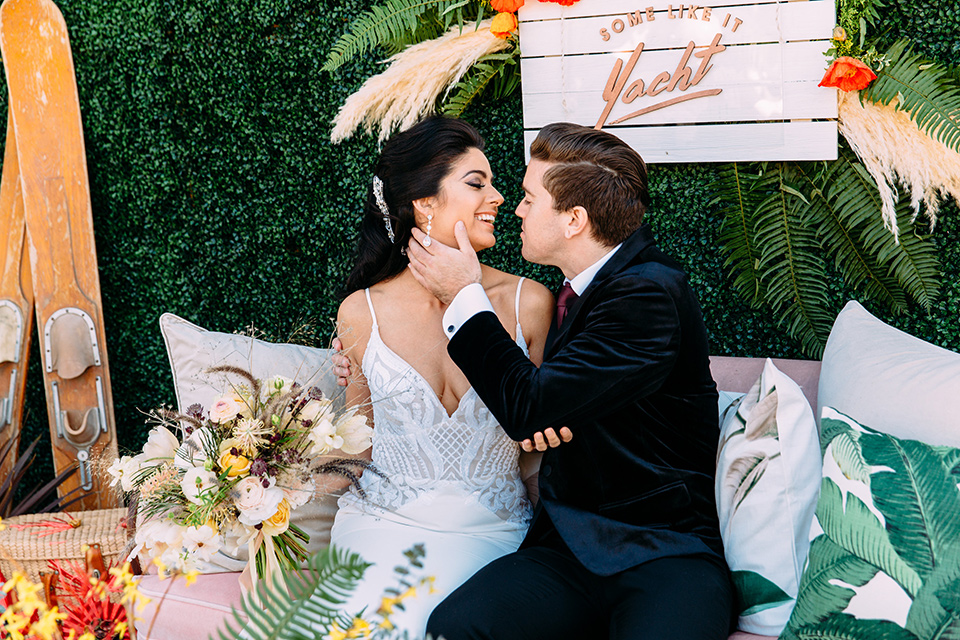 groom touching brides face with a the bride wearing a form fitting  with lace detailing and a deep v neckline the groom is in a tuxedo with peak lapel and burgundy long tie