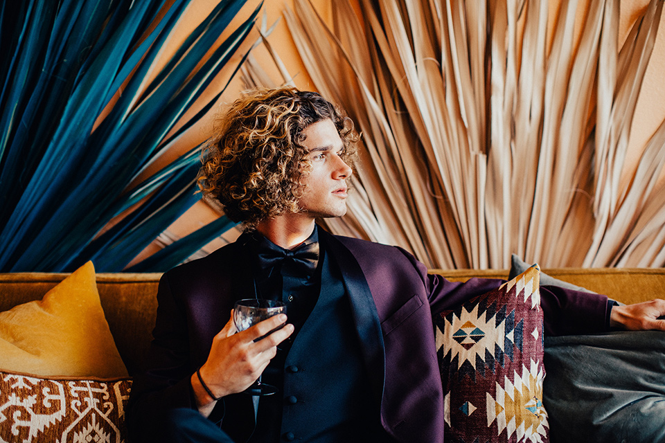 wedding groomsman in a burgundy tuxedo holding a cocktail groomsman is a wearing a deep burgundy tuxedo with a black shirt and black long tie