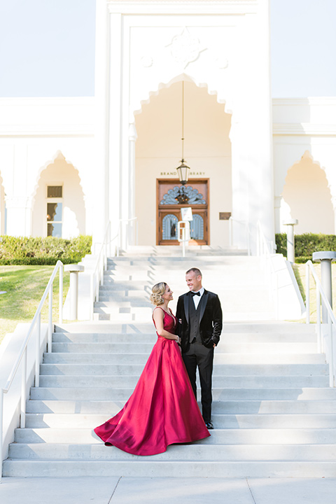  bride in a red evening gown with a plunging neckline and hair in a bun, groom in a black velvet tuxedo and black bow tie 