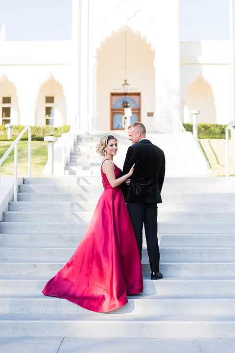 bride in a red evening gown with a plunging neckline and hair in a bun, groom in a black velvet tuxedo and black bow tie