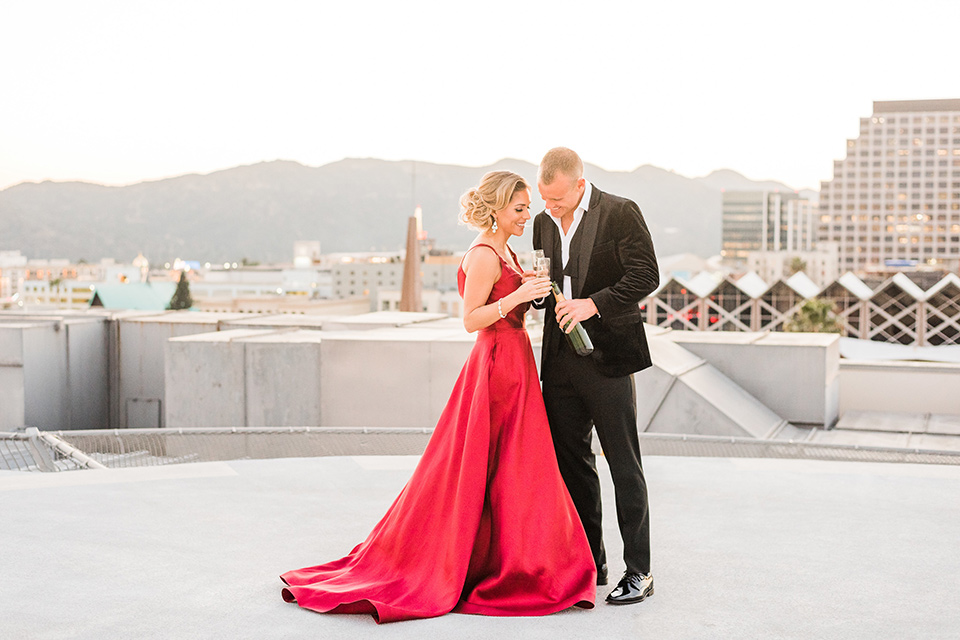  bride in a red evening gown with a plunging neckline and hair in a bun, groom in a black velvet tuxedo and black bow tie 