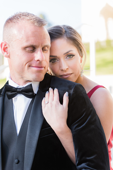 bride in a red evening gown with a plunging neckline and hair in a bun, groom in a black velvet tuxedo and black bow tie