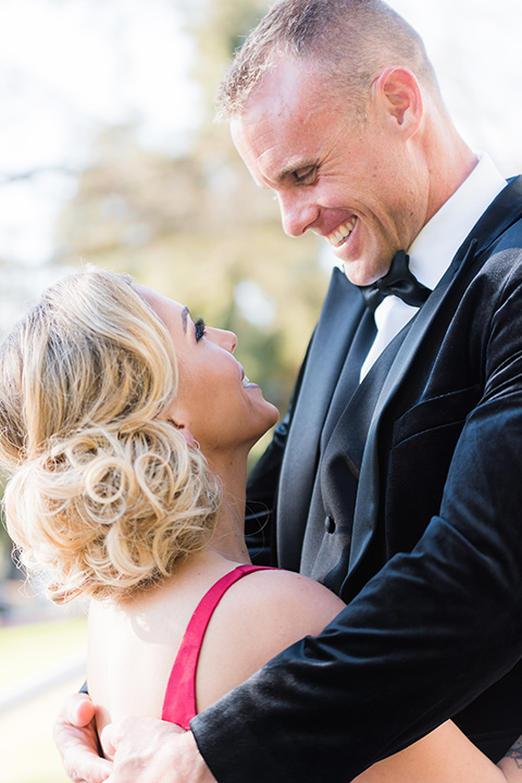  bride in a red evening gown with a plunging neckline and hair in a bun, groom in a black velvet tuxedo and black bow tie 