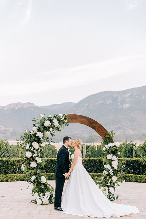 bride and groom by rounded ceremony arch with flowers on it, bride in a white strapless ballgown with a bow detail and hair in a bun, groom in a black tuxedo with a black bow tie