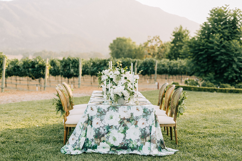  table set up with a floral table linen and wooden chairs with white candles and florals 