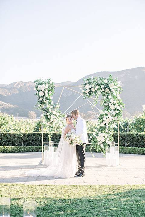 bride and groom at ceremony, bride in a white gown with a plunging neckline and a sheer cape and train and the groom in a white jacket and black pants