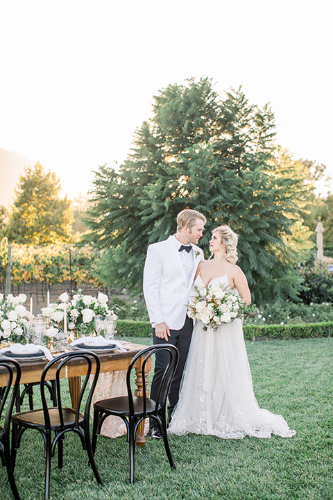 bride and groom at reception table, bride in a white gown with a plunging neckline and a sheer cape and train and the groom in a white jacket and black pants