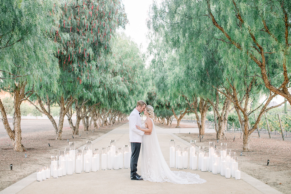  bride and groom by tall candles, bride in a white gown with a plunging neckline and a sheer cape and train and the groom in a white jacket and black pants 