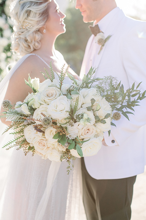  bride and groom close up, bride in a white gown with a plunging neckline and a sheer cape and train with her hair in a bun, groom in a white tuxedo coat and black pants 