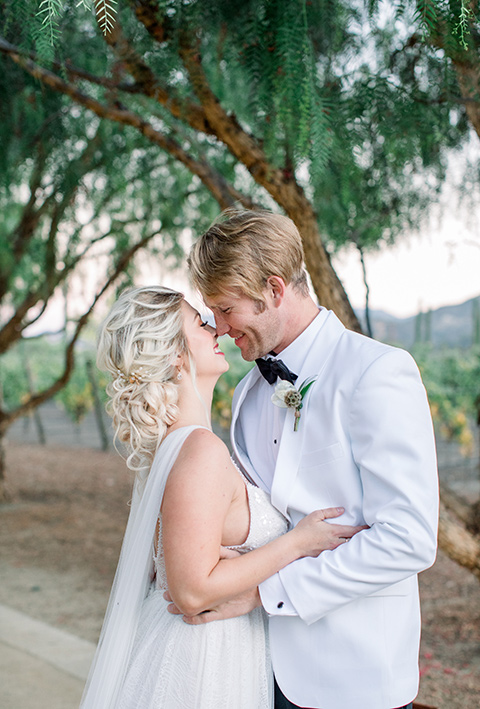 bride and groom close up, bride in a white gown with a plunging neckline and a sheer cape and train and the groom in a white jacket and black pants