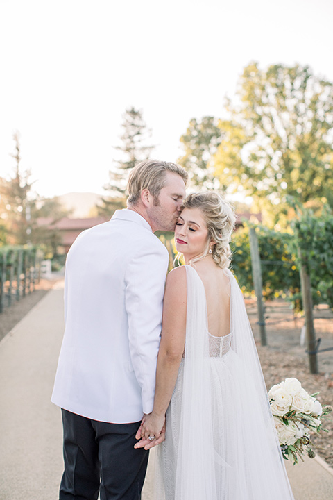  bride in a white gown with a plunging neckline and a sheer cape and train and the groom in a white jacket and black pants