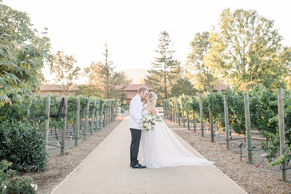 bride in a white gown with a plunging neckline and a sheer cape and train and the groom in a white jacket and black pants 