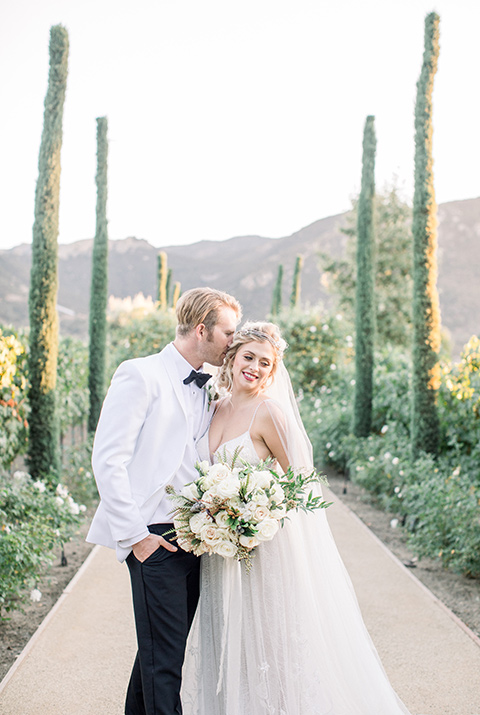 bride and groom by cypress trees, bride in a white gown with a plunging neckline and a sheer cape and train and the groom in a white jacket and black pants 