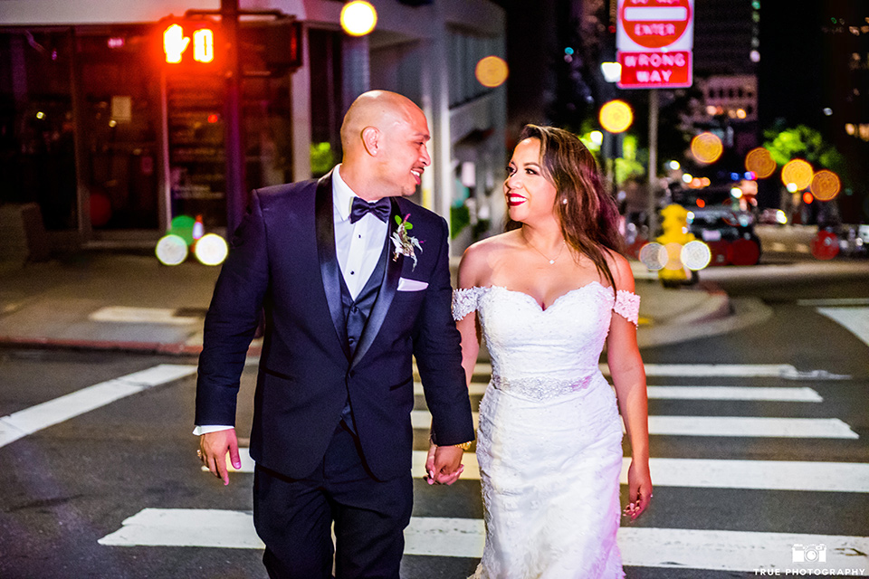 San-Diego-Wedding-bride-and-groom-crossing-street-the-bride-is-in-a-lace-form-fitting-gown-with-an-off-the-shoulder-neckline-groom-is-in-a-black-tuxedo-with-a-black-bow-tie