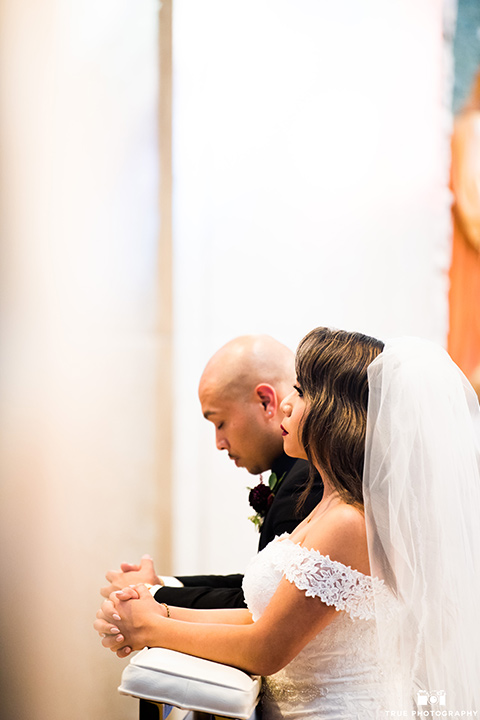 San-Diego-Wedding-bride-and-groom-kneeling-at-ceremony-the-bride-is-in-a-white-lace-gown-with-an-off-the-shoulder-neckline-groom-in-a-black-tuxedo-with-a-black-bow-tie