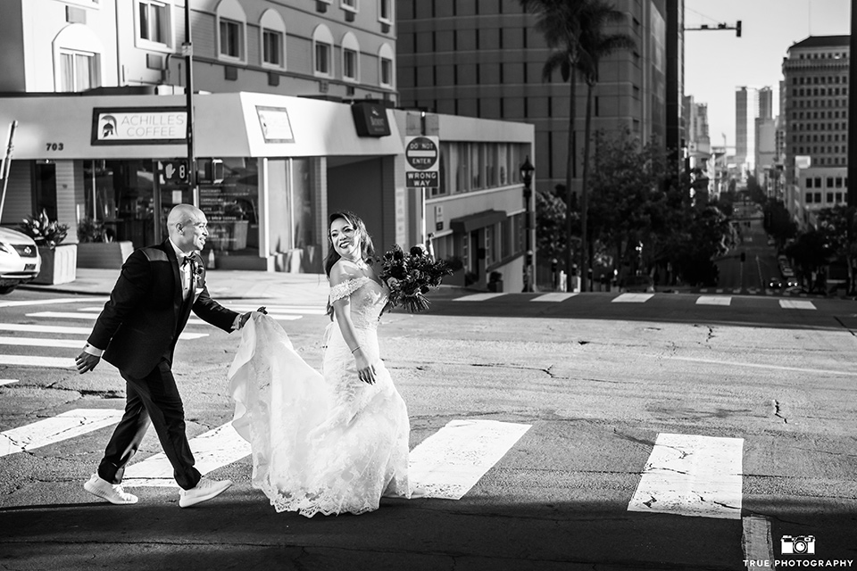 San-Diego-Wedding-groom-holding-brides-gown-crossing-the-street-bride-is-in-a-lace-form-fitting-gown-with-an-off-the-shoulder-neckline-groom-is-in-a-black-tuxedo-with-a-black-bow-tie