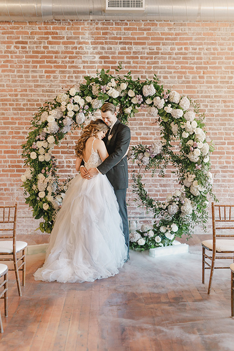 bride in a tulle white gown with a low back detail and a sweetheart neckline and the groom in a black notch lapel tuxedo with a black long tie and a floral circular arch