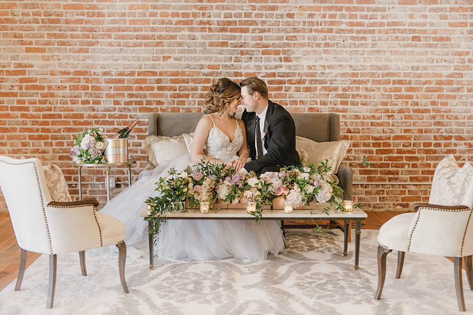 bride in a white tulle ballgown with a low back and a sweetheart neckline and the groom in a black tuxedo with a long black tie sitting on a couch