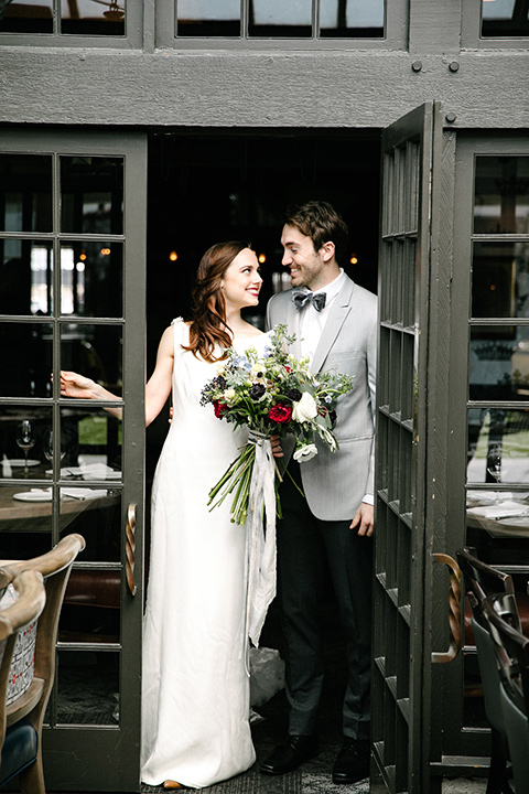 bride in a white lace gown with cap sleeves and a light grey suit coat and black pants with a grey velvet bow tie