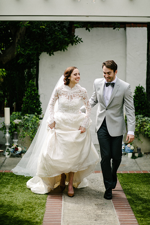 bride in a white flowing gown with long and high neckline sleeves and the groom in a grey suit coat with a pair of black pants and a velvet grey bow tie