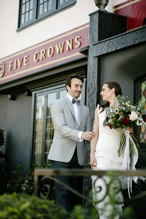 bride in a white flowing gown with long and high neckline sleeves and the groom in a grey suit coat with a pair of black pants and a velvet grey bow tie