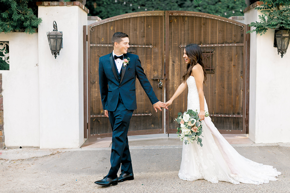 franciscan-gardens-wedding-bride-and-groom-walking-groom-looking-back-at-her-bride-in-a-white-mermaid-strapless-gown-with-hair-down-and-simple-groom-in-a-navy-shawl-lapel-tuxedo-with-a-black-bow-tie