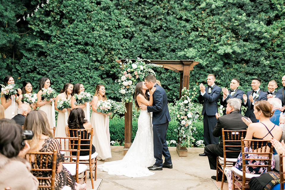 franciscan-gardens-wedding-ceremony-first-kiss-bridesmaids-in-a-light-chamagne-color-groomsmen-in-navy-suits-bride-in-a-white-mermaid-strapless-gown-with-hair-down-and-simple-groom-in-a-navy-shawl-lapel-tuxedo-with-a-black-bow-tie
