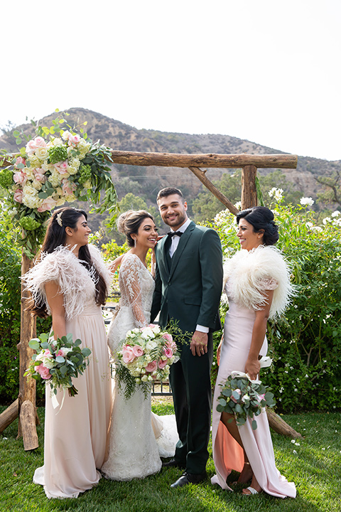 bride in a lace gown with long sleeves and a bouquet of white and pink florals bridesmaids in light pink gowns and the groom in a dark green suit