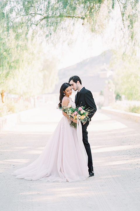 bride in a blush colored gown with thin straps and a sweetheart neckline and the groom in a black shawl lapel tuxedo with a black long tie