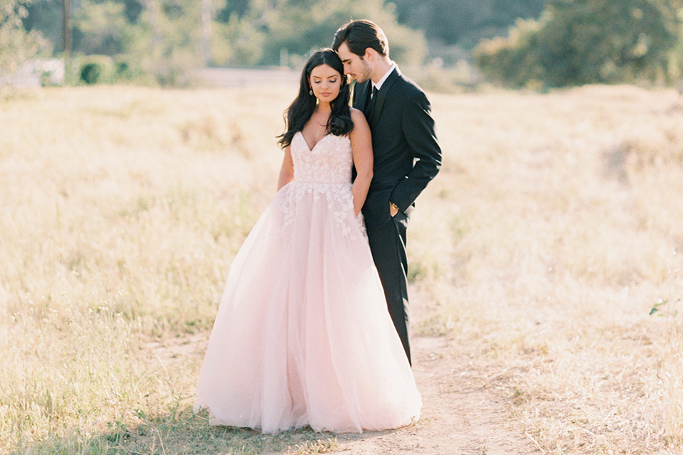 bride in a blush tulle ballgown with a low back and a sweetheart neckline and the groom in a black tuxedo with a long black tie sitting on a couch