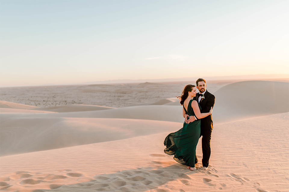 Glamis-Engagement-Shoot-bride-and-groom-holding-eachother-bride-in-a-deep-green-flowing-dress-with-a-plunging-neckline-groom-in-a-black-tuxedo-with-navy-blue-velvet-bow-tie