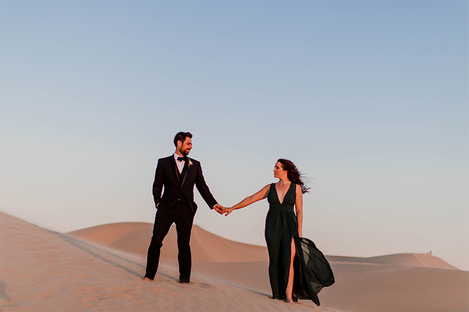 Glamis-Engagement-Shoot-bride-and-groom-holding-hands-bride-in-a-deep-green-flowing-dress-with-a-plunging-neckline-groom-in-a-black-tuxedo-with-navy-blue-velvet-bow-tie
