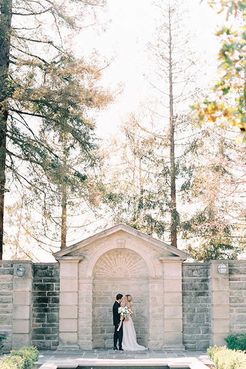 bride wearing a tulle ballgown with thin straps and her hair in a loose braided bun and the groom in a black tuxedo with a black bow tie in front of a brick building