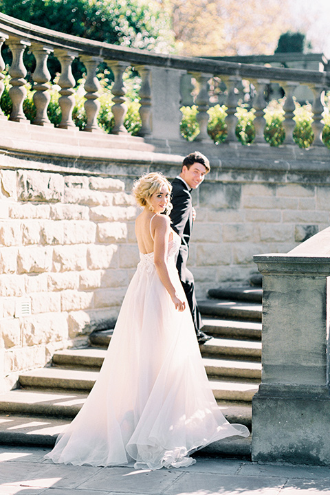  bride wearing a tulle ballgown with thin straps and her hair in a loose braided bun with a peony and rose bouquet and the groom in a black tuxedo and a black bow tie walking up the stairs