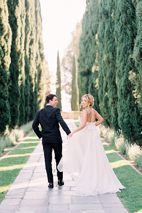  bride wearing a tulle ballgown with thin straps and her hair in a loose braided bun with a peony and rose bouquet and the groom in a black tuxedo and a black bow tie walking away