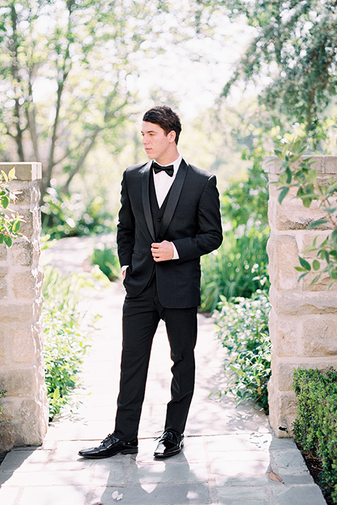  the groom in a black tuxedo with a black bow tie standing by archway