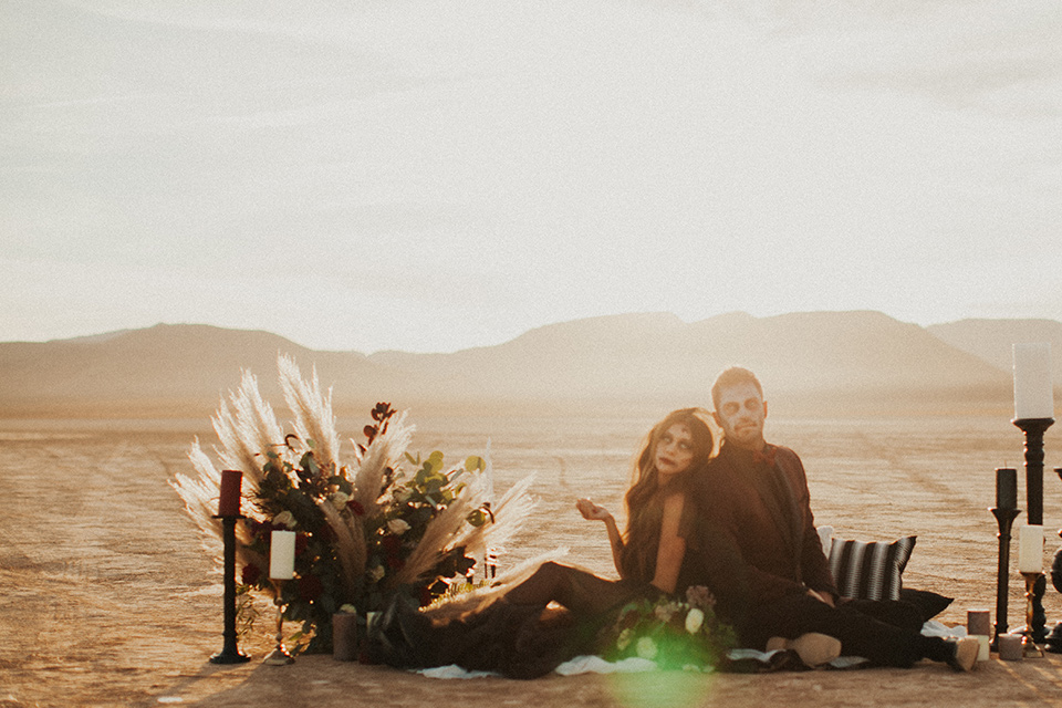 halloween shoot with the bride in a black and burgundy tulle gown with a black veil and skeleton makeup and the groom in a burgundy tuxedo with a black shirt and burgundy bow tie sitting on a carpet in the desert with candles and florals