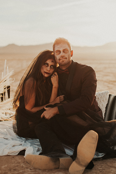 Halloween shoot with the bride in a black and burgundy tulle gown with a black veil and the groom in a burgundy tuxedo with a black trim, a black shirt, and a burgundy bow tie sitting down