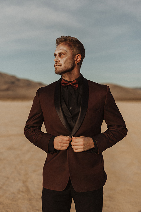 Halloween shoot with the groom in a burgundy tuxedo with a black trim, a black shirt, and a burgundy bow tie sitting down