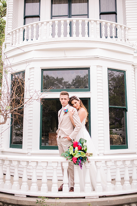  bride in a boho style gown with a bright colored floral crown and the groom in a tan suit with a tan long tie by a white building