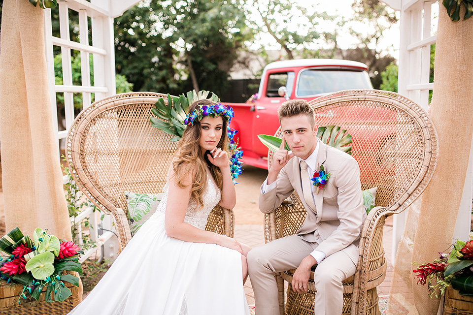 bride in a boho style gown with a bright colored floral crown and the groom in a tan suit with a tan long tie sitting in big chairs