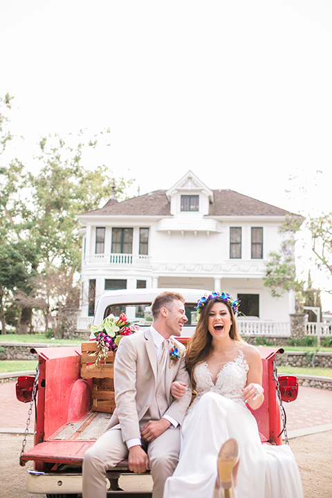  bride in a boho style gown with a bright colored floral crown and the groom in a tan suit with a tan long tie in the bed of a truck