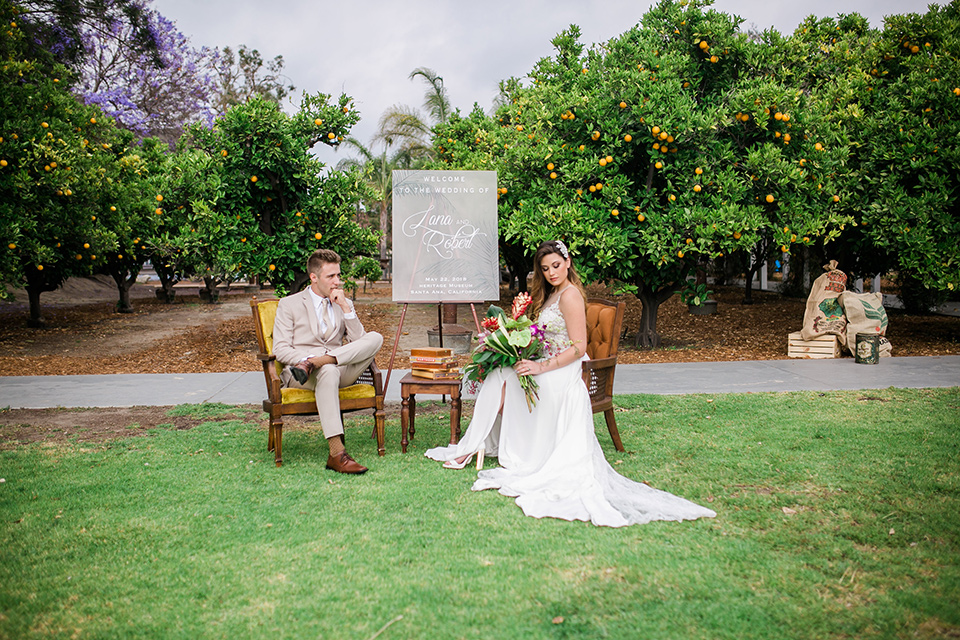 bride in a boho style gown with a bright colored floral crown and the groom in a tan suit with a tan long tie sitting in chairs outside on the grass